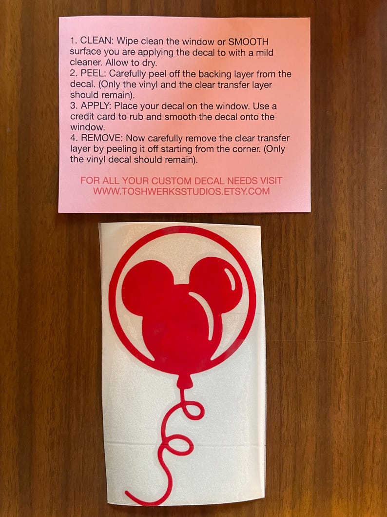 May include: A red vinyl decal featuring a Mickey Mouse balloon design. The decal is on a white backing, with instructions on a pink paper above. The instructions detail how to apply the decal to a window or smooth surface.