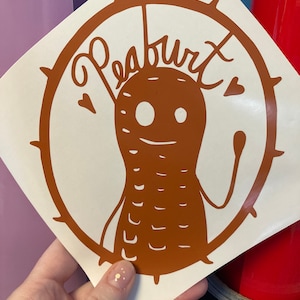 May include: A white sticker with a brown peanut-shaped character with a smiling face and the word "Peaburt" written above it. The character is surrounded by a circle with spikes.
