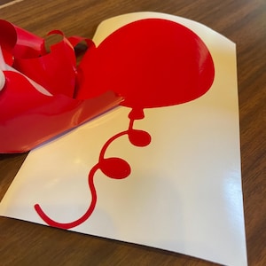 May include: A red balloon sticker with a curved string and three small circles. The balloon is on a white background, with red backing material partially peeled away. The sticker is likely for decoration.