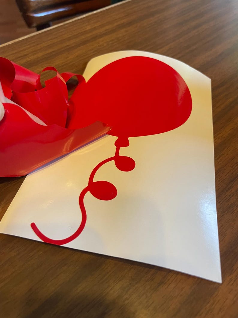 May include: A red balloon sticker with a curved string and three small circles. The balloon is on a white background, with red backing material partially peeled away. The sticker is likely for decoration.