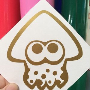 Könnte beinhalten: Goldener Vinyl-Aufkleber mit einem Cartoon-Tintenfisch mit großen Augen und einem gepunkteten Körper. Der Aufkleber befindet sich auf einem weißen Hintergrund.