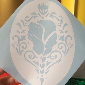 May include: A white vinyl stencil with a decorative floral design featuring a stylized rose in the center. The stencil is on a light blue background.