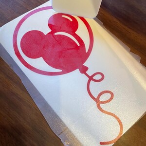 May include: A red Mickey Mouse balloon vinyl decal being applied with a white squeegee. The balloon design is inside a circle, with a curly ribbon. The decal is on a white backing.