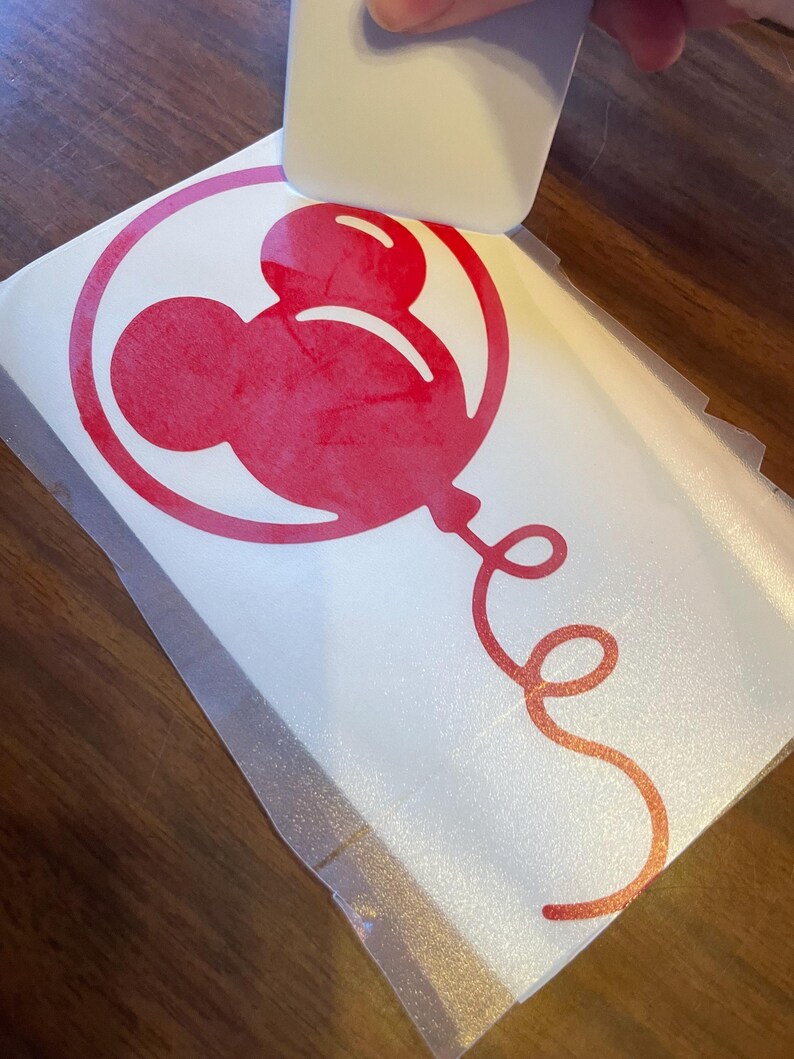 May include: A red Mickey Mouse balloon vinyl decal being applied with a white squeegee. The balloon design is inside a circle, with a curly ribbon. The decal is on a white backing.