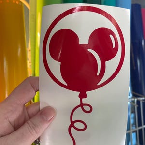 May include: A red vinyl decal featuring a Mickey Mouse balloon design. The balloon is a stylized silhouette with a round shape and two smaller circles for ears, enclosed in a red circle. A curly red string hangs below.