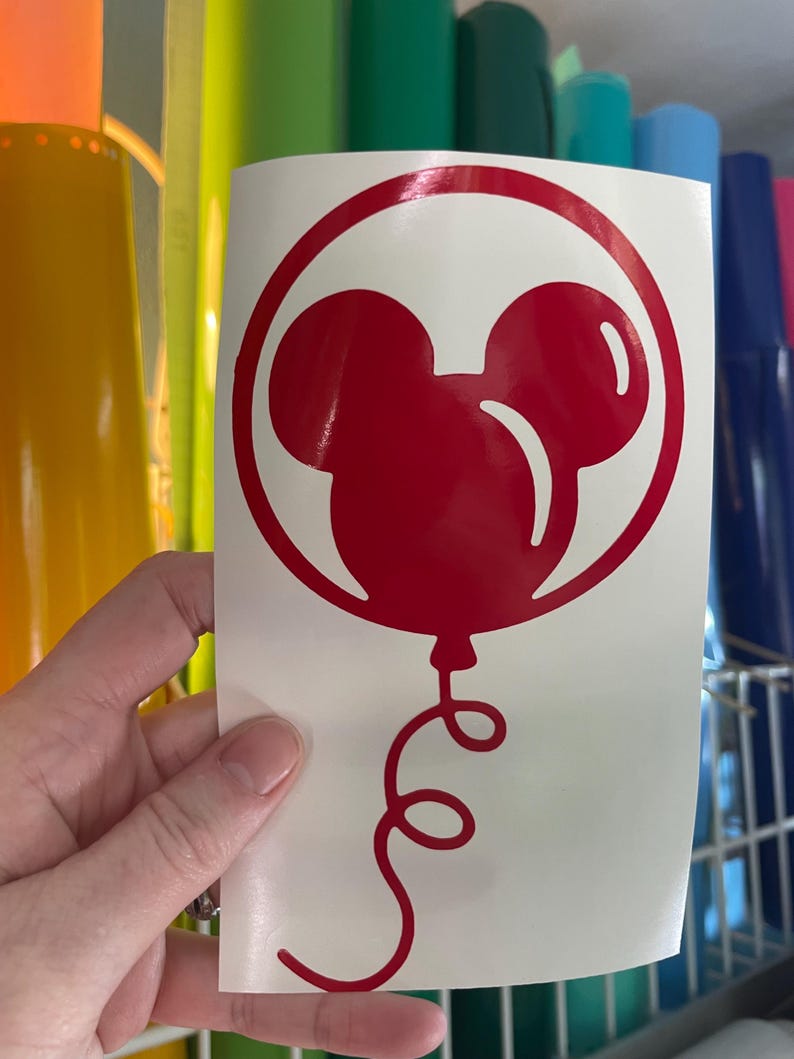 May include: A red vinyl decal featuring a Mickey Mouse balloon design. The balloon is a stylized silhouette with a round shape and two smaller circles for ears, enclosed in a red circle. A curly red string hangs below.