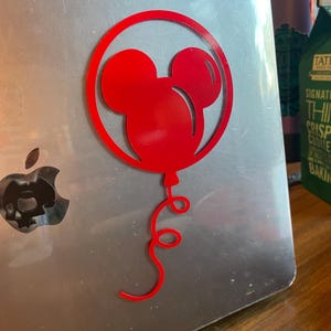 May include: A red Mickey Mouse balloon sticker on a silver laptop. The sticker features the iconic mouse head within a circular frame, with a curly balloon string. The laptop has a skull-shaped design in place of the Apple logo.