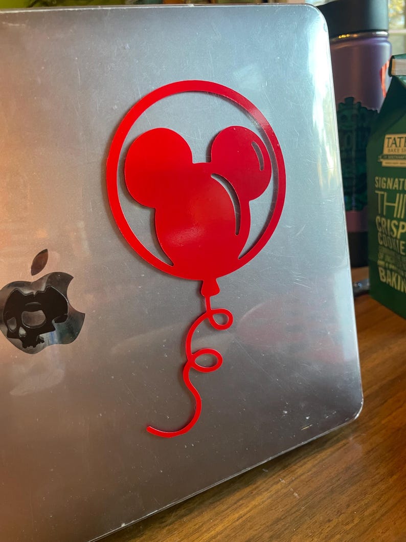 May include: A red Mickey Mouse balloon sticker on a silver laptop. The sticker features the iconic mouse head within a circular frame, with a curly balloon string. The laptop has a skull-shaped design in place of the Apple logo.