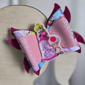May include: A large, decorative hair bow featuring a pink glitter center and magenta glitter accents. The bow has a cartoon print with Princess Peach and Toad from the Super Mario Bros. video game series. The bow is set against a light wooden background.