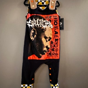 May include: Black overalls with a graphic print of a person's face with a crown of thorns. The overalls have orange buttons and checkerboard print on the legs with yellow smiley faces.