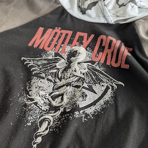 以下が含まれることがあります： 赤色の文字で「Mötley Crüe」というバンド名が入った黒いパーカー。グラフィックは、ヘビのような尾を持つ翼のある生き物とスターバーストのデザインが特徴です。フードの裏地はコウモリとクモの巣模様です。