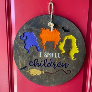 May include: A round wooden sign with three witches' faces, a broom, and bats. The sign says "I smell children".
