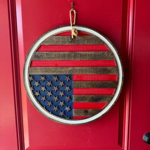 May include: A round wooden American flag wall hanging. The flag design features red and brown stripes, a blue field with stars, and a white border. It is suspended by a jute rope from a metal hook on a red door.