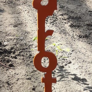 May include: An orange metal garden stake with the word "Carrots" cut out. The stake is placed in the soil, with small plants visible in the background. The stake is approximately 12 inches tall.
