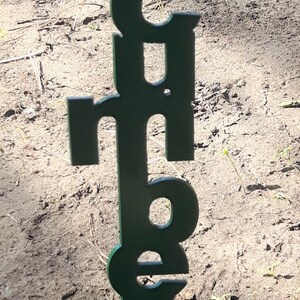 May include: A dark green metal garden stake with the word "Cucumbers" cut out. The stake is placed in the soil, with the letters vertically aligned. The background is soil and small plants.