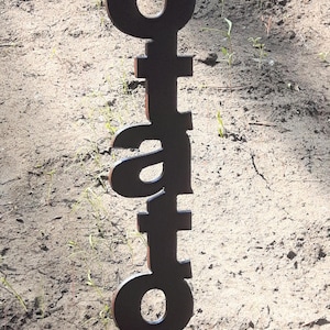 May include: A dark brown metal garden stake with the word "Potatoes" cut out in a vertical orientation. The stake is placed in the soil, with small green plants visible in the background.