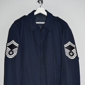 Op de afbeelding: Een donkerblauwe bomberjack met een ritssluiting en twee witte en zwarte patches op de mouwen. De patches hebben drie zilveren strepen en een ster.