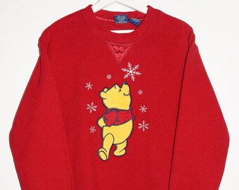 Op de afbeelding: Rode longsleeve trui met een Winnie de Poeh-ontwerp. De beer is geel en draagt een rode sjaal. Het ontwerp bevat sneeuwvlokken en de tekst "Winnie de Poeh".