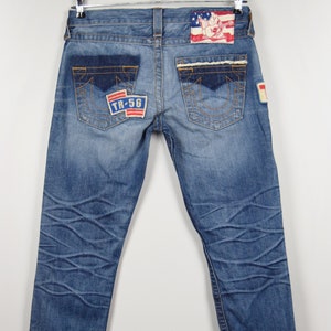 Puede incluir: Vaqueros de mezclilla azul con detalles desgastados y parches. Los parches presentan una bandera estadounidense roja, blanca y azul, un logotipo rojo, blanco y azul con el texto "TR-56" y un parche rojo, blanco y azul con una figura de dibujos animados.