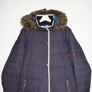 May include: A dark blue puffer jacket with a faux fur hood and a white zipper trim. The jacket has two zippered pockets on the front and a zippered pocket on the left sleeve. The jacket has a brown leather patch with the word "Joules" on the right sleeve.