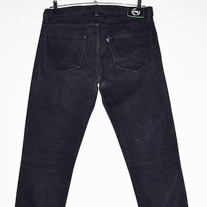 May include: Dark wash denim jeans hanging on a metal hanger. The jeans have a classic five-pocket design, with two back pockets and a small coin pocket. The jeans are a deep black color and appear to be made of a durable denim material.