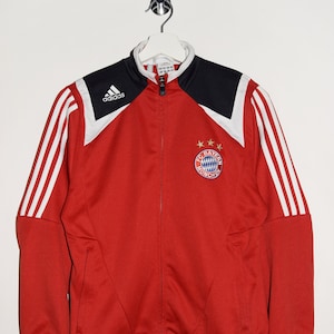 May include: Red Adidas track jacket with white stripes and a black panel on the shoulders. The jacket has a zipper closure and a Bayern Munich logo on the chest.
