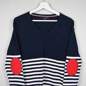 Puede incluir: Un suéter de manga larga a rayas azul marino y blanco con parches rojos en los codos. El suéter tiene cuello en V y el nombre de la marca "Tommy Hilfiger" es visible en la etiqueta.