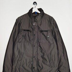Könnte beinhalten: Eine dunkelgraue, leichte Jacke mit Reißverschluss und Knopfleiste. Die Jacke hat mehrere Taschen und einen Stehkragen.