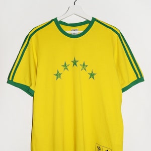 May include: Yellow short-sleeved t-shirt with green stripes on the sleeves and a green trim around the neck. The shirt has five green stars on the front and a black Adidas logo on the bottom right.