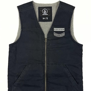 May include: A navy blue vest with a gray fleece lining and a full-length zipper. The vest features two front pockets and two patches with the "VOLCOM" logo and "ANTI HERO" text. The vest is designed for outerwear.