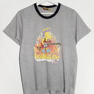 Könnte beinhalten: Ein graues T-Shirt mit einem schwarzen gerippten Rundhalsausschnitt. Die Vorderseite des Shirts zeigt ein Cartoon-Bild von Bart Simpson, der Gitarre spielt, mit Flammen dahinter. Der Text "Born to Rock" ist unter dem Bild gedruckt. Der Text "The Simpsons" ist unter dem Text "Born to Rock" gedruckt.
