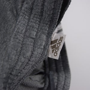 Puede incluir: Primer plano de una etiqueta Adidas gris cosida en una prenda gris. La etiqueta tiene el logotipo de Adidas y la palabra "adidas" impresa en negro.