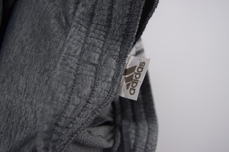 Puede incluir: Primer plano de una etiqueta Adidas gris cosida en una prenda gris. La etiqueta tiene el logotipo de Adidas y la palabra "adidas" impresa en negro.