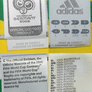 May include: A yellow soccer jersey with the FIFA World Cup Germany 2006 logo and the Adidas logo. The jersey has a white tag with the text "Official Licensed Product" and a white tag with size information in multiple languages. The jersey also has a white tag with the text "The Official Emblem, the Official Mascots of the 2006 FIFA World Cup Germany" and the text "FIFA World Cup Trophy are copyrights and trademarks of FIFA. All rights reserved. Manufactured under licence by Adidas."