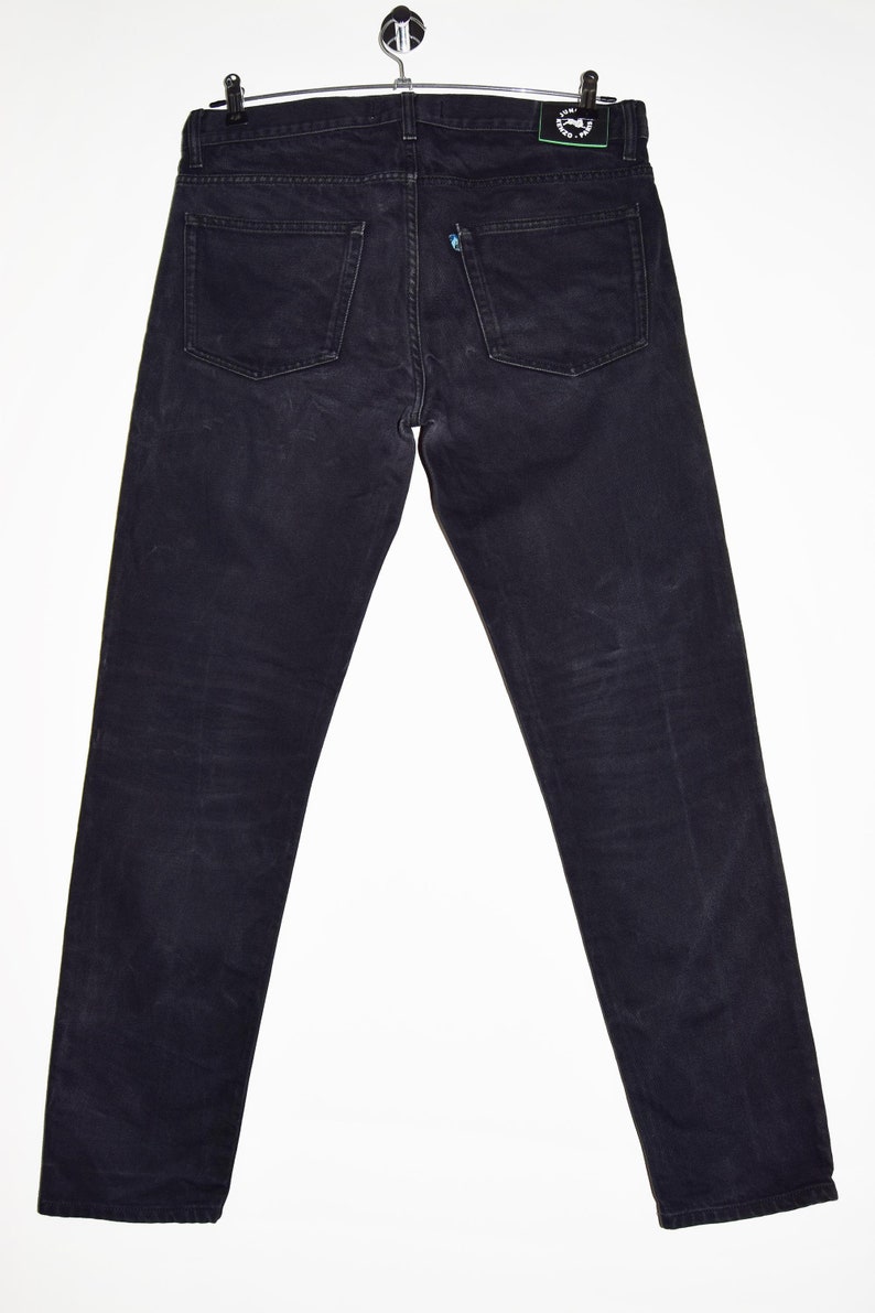 May include: Dark wash denim jeans hanging on a metal hanger. The jeans have a classic five-pocket design, with two back pockets and a small coin pocket. The jeans are a deep black color and appear to be made of a durable denim material.