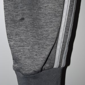 Puede incluir: Pantalones de ch&aacute;ndal grises con una raya blanca en cada pierna. Los pantalones de ch&aacute;ndal tienen un pu&ntilde;o de canal&eacute; en la parte inferior.