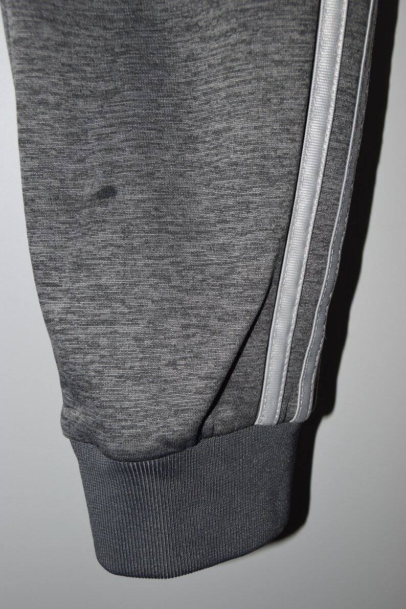 Puede incluir: Pantalones de ch&aacute;ndal grises con una raya blanca en cada pierna. Los pantalones de ch&aacute;ndal tienen un pu&ntilde;o de canal&eacute; en la parte inferior.