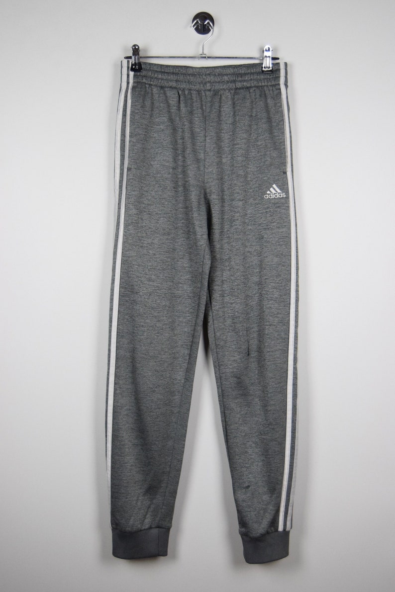 Puede incluir: Pantalones de ch&aacute;ndal grises con rayas blancas a los lados y un logotipo de Adidas blanco en la pierna izquierda.