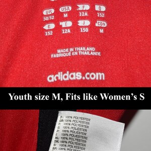 May include: Red Adidas youth size shirt with white text and size tags. The text reads "adidas", "Youth size M, Fits like Women's S", "Made in Thailand", "Fabriqu&eacute; en Tha&iuml;lande", and "adidas.com". The size tags list sizes in UK, USA, F, I, J, and CDN.