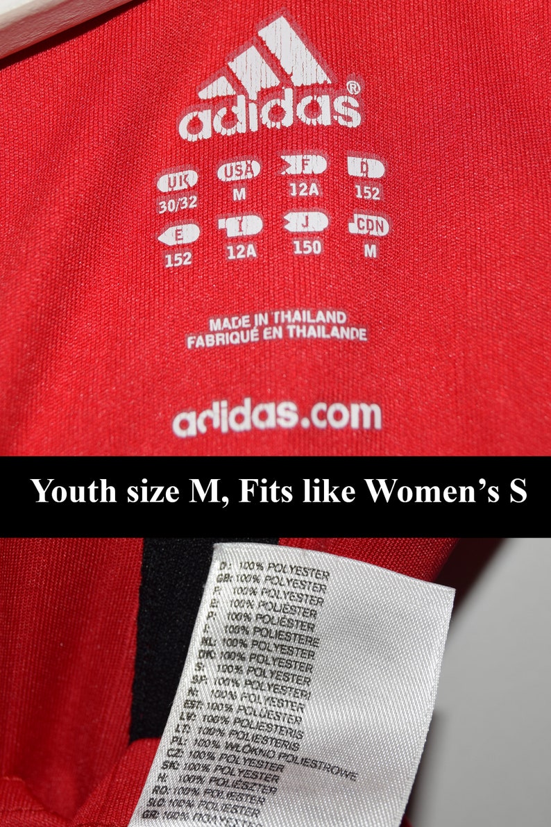 May include: Red Adidas youth size shirt with white text and size tags. The text reads "adidas", "Youth size M, Fits like Women's S", "Made in Thailand", "Fabriqu&eacute; en Tha&iuml;lande", and "adidas.com". The size tags list sizes in UK, USA, F, I, J, and CDN.