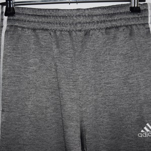 Puede incluir: Pantalones de ch&aacute;ndal grises con rayas blancas a los lados y el logotipo de Adidas en la pierna derecha.