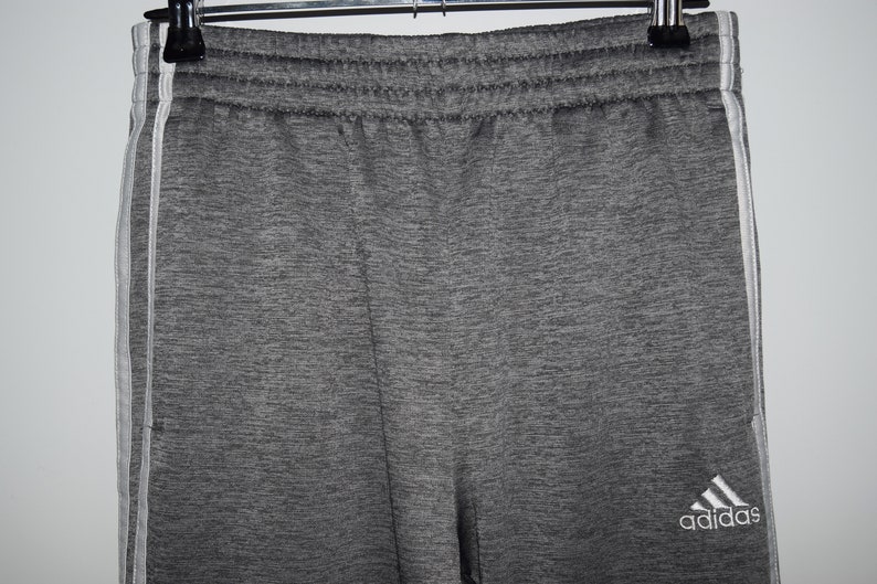 Puede incluir: Pantalones de ch&aacute;ndal grises con rayas blancas a los lados y el logotipo de Adidas en la pierna derecha.
