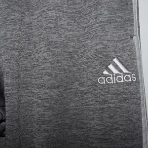 Puede incluir: Pantalones de ch&aacute;ndal grises con un logotipo de Adidas blanco en la pierna derecha.