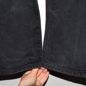 May include: Close-up of black denim pants. The pants are made of a dark, textured fabric. The hem of the pants is visible, with a stitched edge. The pants are slightly worn, with some fading.
