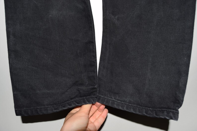 May include: Close-up of black denim pants. The pants are made of a dark, textured fabric. The hem of the pants is visible, with a stitched edge. The pants are slightly worn, with some fading.