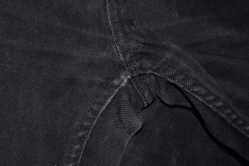 May include: Close-up of black denim fabric, showing the texture and stitching details. The image highlights the seams and construction of the garment, with a focus on the durable material and craftsmanship.