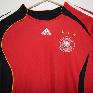 May include: Red soccer jersey with black and yellow accents. The Adidas logo is on the chest, along with the German national team crest and three stars.