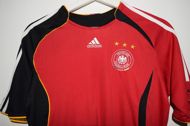 May include: Red soccer jersey with black and yellow accents. The Adidas logo is on the chest, along with the German national team crest and three stars.