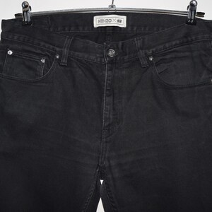 May include: Black denim jeans hanging on a hanger. The jeans have a classic five-pocket design with a button and zipper closure. The label reads "KENZO x H&M".