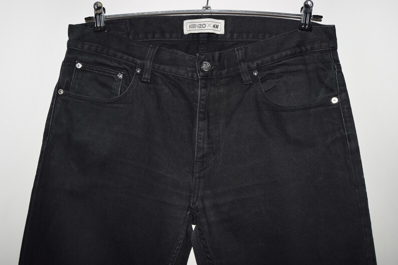 May include: Black denim jeans hanging on a hanger. The jeans have a classic five-pocket design with a button and zipper closure. The label reads "KENZO x H&M".